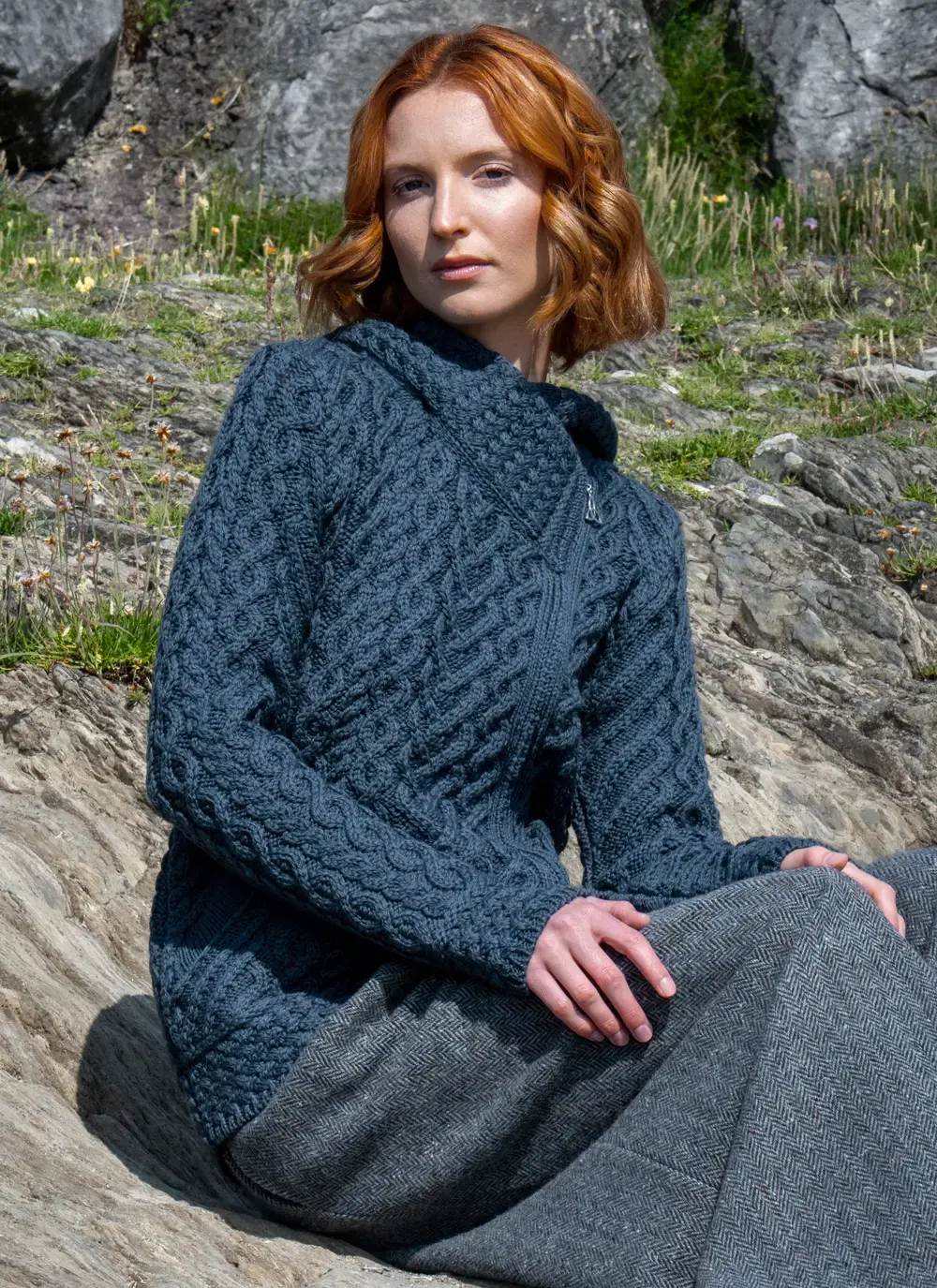 Red headed model sitting in rocky area wearing a blue Aran biker hoodie